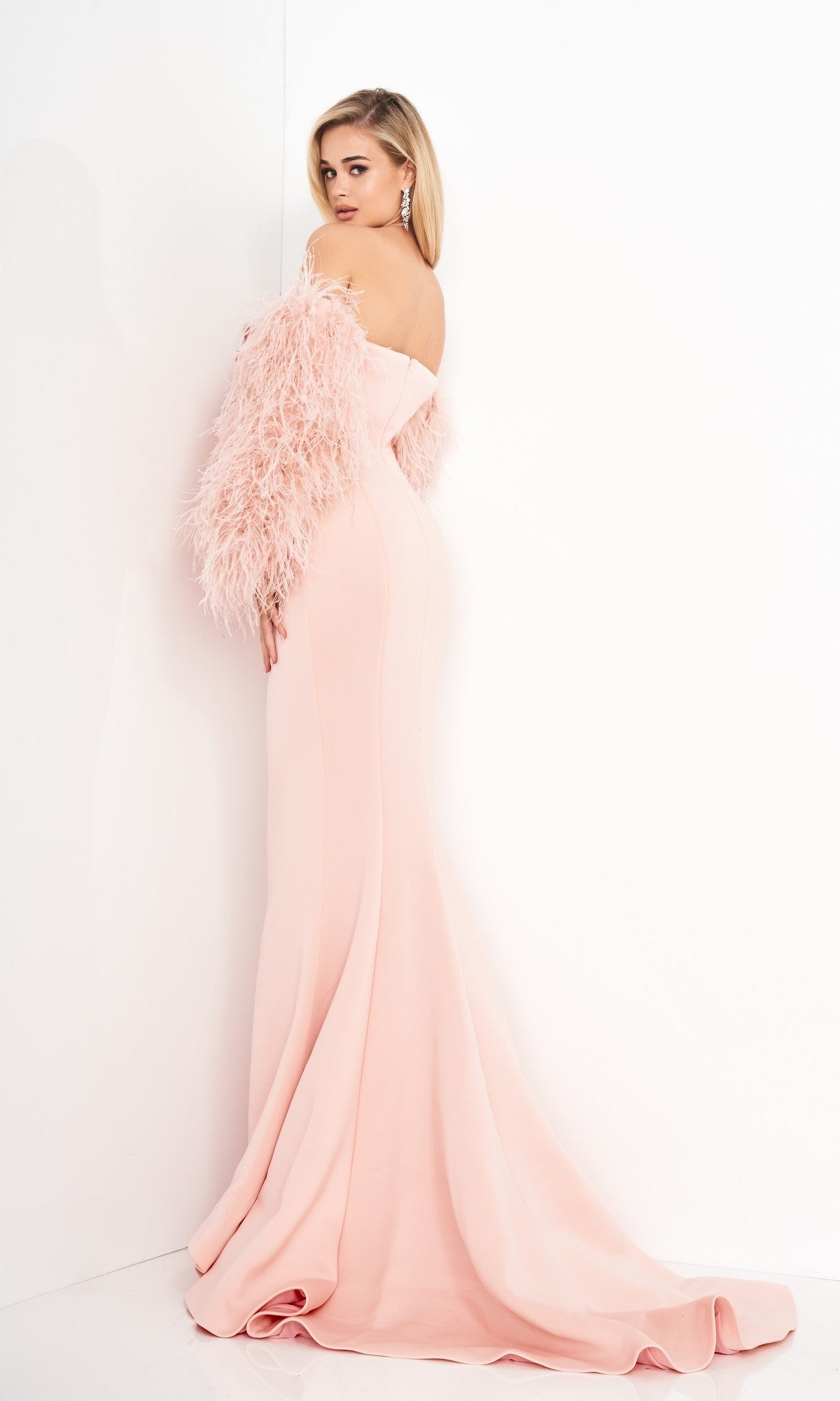 A woman in the Jovani 1226 long prom dress with feather sleeves stands against a white wall, looking over her shoulder.